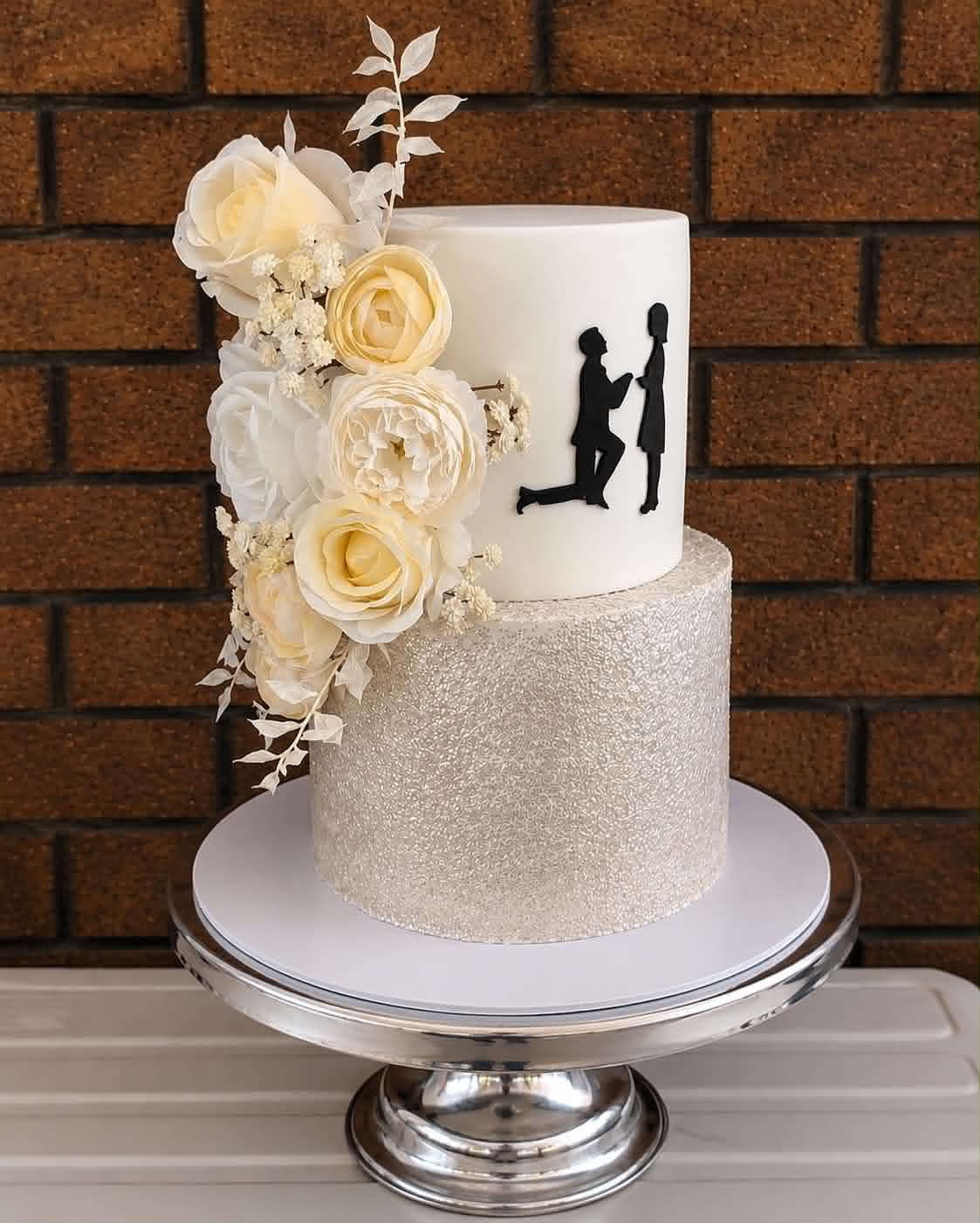Two tier engagement cake with silver glitter bottom tier, white fondant top tier with black proposal silhouette, cascading white and cream roses, ranunculus and gypsophila